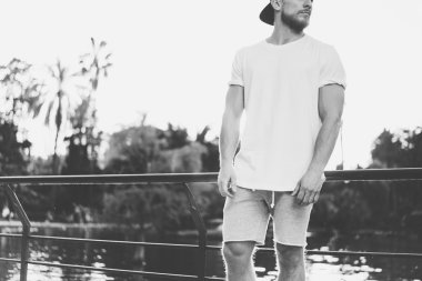 Görüntü Sakallı Kaslı Adam Beyaz Boş t-shirt, snapback kap ve şort yaz tatilinde giyiyor. Göl kenarında dinlendirici bir zaman. Green City Garden Park Sunset Background.Back görünümü. Yatay Mockup