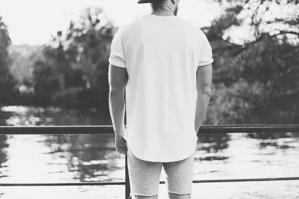 Foto Bearded muskulös man klädd i blank t-shirt, snapback mössa och shorts på sommaren tid. Avkopplande tid nära sjön. Green City Garden Park Sunset bakgrund. tillbaka vy. Svart vit mockup — Stockfoto