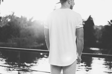 Resim Sakallı Kaslı Adam Boş t-shirt, snapback kap ve şort yaz tatilinde giyiyor. Göl kenarında dinlendirici bir zaman. Green City Garden Park Sunset Background.Back görünümü. Siyah Beyaz Mocku
