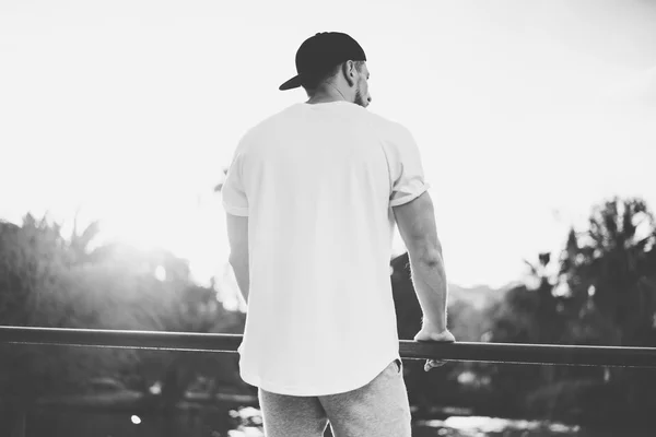 Foto hombre musculoso barbudo con camiseta en blanco, gorra de snapback y pantalones cortos en verano. Green City Garden Park Sunset Background (en inglés). Vista trasera. Mockup.Black horizontal blanco — Foto de Stock