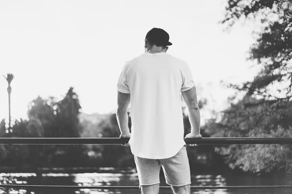 Bearded Muscular Man Wearing Blank t-shirt, snapback cap and shorts in summer time.Relaxing  near the lake.Green City Garden Park Sunset Background. Back view. Horizontal Mockup.Black White — Stock Photo, Image