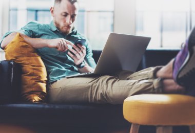 Sakallı Hipster çalışma Laptop modern Tasarım İç Studio Loft.Men Relax Vintage Kanepe, Çağdaş Notebook kullanın, Smartphone.Blurred Background.Creative Business Startup Idea.Film, parlama