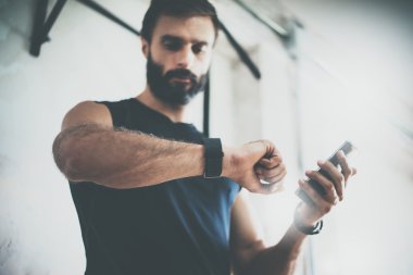 Fotoğraf sakallı sportif adam sonra egzersiz oturum denetler Fitness sonuçları Smartphone.Adult adam giyiyor spor Tracker bileklik Arm.Training spor salonu içinde zor. Yatay bar arka plan. Bulanık.