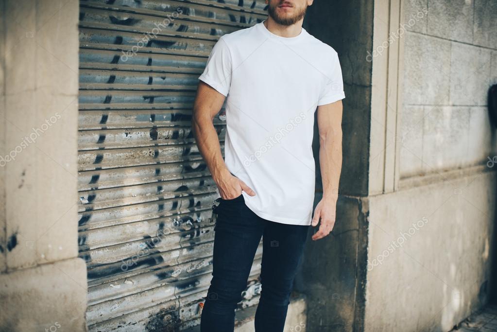 Portrait of a bearded man wearing blank t-shirt, blue jeans and standing on the street next to the garage