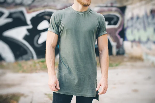 Skäggig kille klädd i grön blank t-shirt och svarta jeans, stående på gatan — Stockfoto