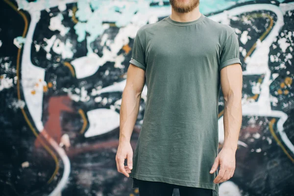 Skäggig kille klädd i grön blank t-shirt och svarta jeans, stående på gatan. horisontella — Stockfoto