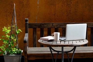 Evden dışarı çalışma kavramı. Laptop, bir fincan kahve, defter ve cep telefonu ahşap kafe masasında. Tahta bir bank ya da yanında yeşil bir bitki olan bir kanepe, turuncu bir binanın önünde..