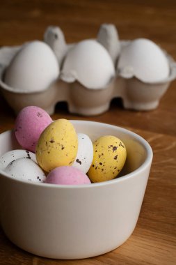 Tahta masadaki bir kasede çeşitli renklerde çikolatalı Paskalya yumurtaları. Arka planda bir yumurta kutusu var..