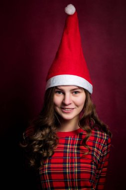 Kıvırcık saçlı, Noel Baba şapkalı, kafesli bir kız kırmızı bir duvara bakıp gülümsüyor ve kameraya bakıyor..