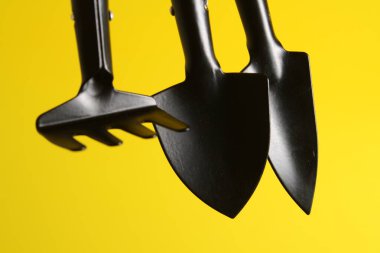 Sarı zemin üzerinde toprak ve çiçeklerle çalışmak için üç küçük nesne, 2 kürek ve tırmık.