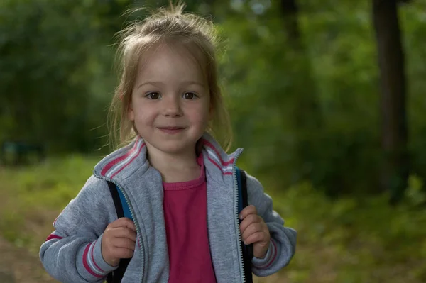 Une petite fille heureuse avec un sac à dos se tient dans un parc forestier de printemps et sourit — Photo