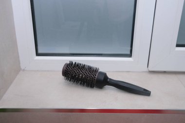 Yuvarlak bir saç fırçası pencere pervazında durur, güzellik rutinlerini ve özbakımını ima eder. Mekan, güzellik ve yaşam tarzı kavramları için ideal bir spa gününü çağrıştırıyor.