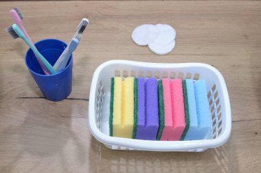 Düzgün ev eşyaları dizisi: sepetteki renkli süngerler, mavi bardaktaki diş fırçaları ve ahşap bir yüzeyde pamuk yastıklar. Odak noktamız temizlik ve hijyen.