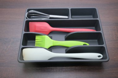 Koyu gri bir kap düzenleyici tepsisi ahşap bir yüzeyde mutfak aletlerini düzgünce düzenliyor. Bir çırpma, spatula ve fırça özelliği var. Ev düzenleme kavramları için mükemmel.