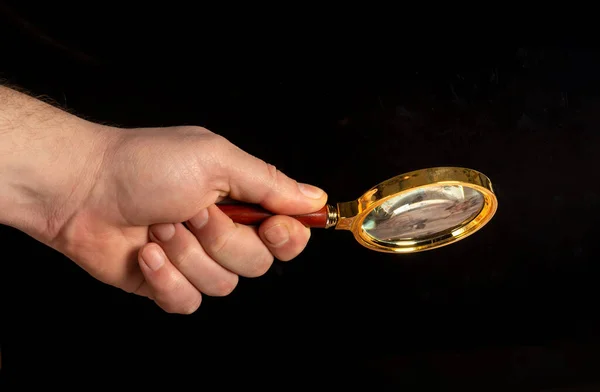 Man hand holding a gilded magnifying glass, close-up on black background. Copy space for your image or text