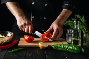 Şefin eli bıçaklı, salata için taze kırmızı domates kesiyor. Restoranın mutfağında çalışma ortamı. Masada taze sebze ve yağ var.
