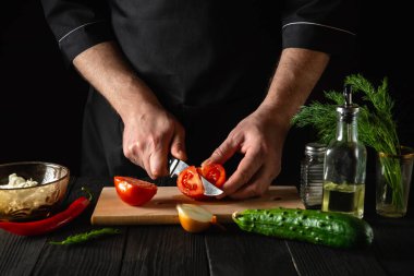 Şefin eli bıçaklı, salata için taze kırmızı domates kesiyor. Restoranın mutfağında çalışma ortamı. Masada taze sebze ve yağ var.