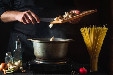 Şef sebze ve spagetti arka planında çorba hazırlıyor. Bir restoranın mutfağındaki tencereye mantar döken bir elin yakın çekimi.