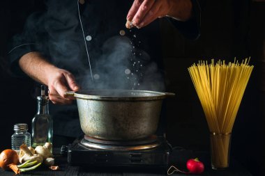 Şef sebzelerin arka planında İtalyan makarnası hazırlıyor. Lokanta mutfağında bir tencereye biber döken bir aşçının yakın çekimi.