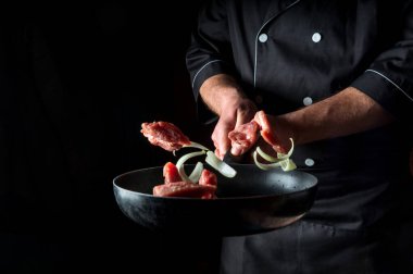 Profesyonel bir aşçının eli siyah arka planda soğanlı et parçalarını kızartma tavasına atar. Restoran pişirme konsepti