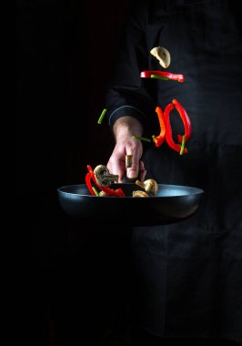 Profesyonel bir aşçının eli siyah arka planda bir tavaya sebze ve mantar parçaları atar. Restoran yemekleri konsepti. Ücretsiz reklam alanı