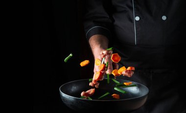 Profesyonel bir aşçının eli siyah arka planda sebze ve et parçalarını kızartma tavasına atar. Restoran yemekleri konsepti. Ücretsiz reklam alanı