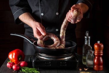 Aşçı pişirmeden önce tavaya çiğ et sosisi koyar. Restoran mutfağında çalışma ortamı, sebzeler ve baharatlarla birlikte antika bir masa.