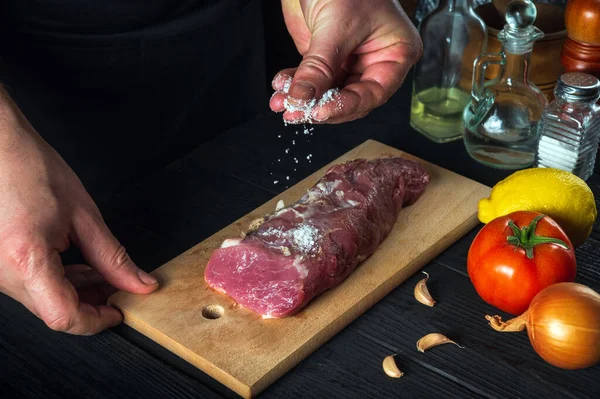 Chef or cook sprinkles the raw beef meat with salt. Preparing meat before baking. Working environment in the kitchen of restaurant or cafe