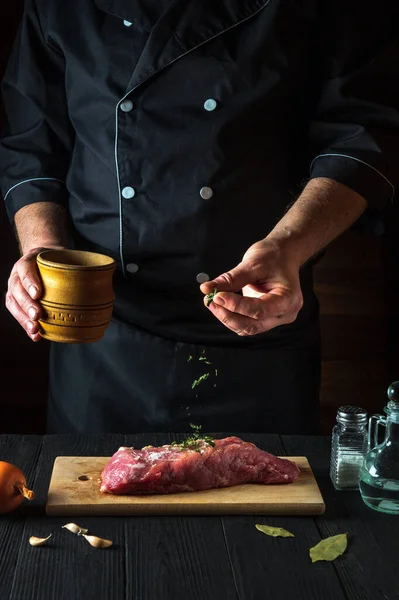 The chef prepares raw beef meat fillets and adds dry spices for the marinade. Working environment in the kitchen in a restaurant or cafe