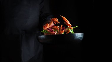 Profesyonel şef sebzeli tavada karides pişirir. Deniz ürünleri pişirmek, sağlıklı vejetaryen yiyecekler ve karanlık bir arka planda yemek. Hareket halindeyken donuyorum. Ücretsiz reklam alanı