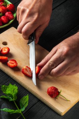 Close-up chef hands cut fresh strawberries on cutting board of restaurant kitchen for making soft drink with mint. Cooking diet desserts. Vertical image.