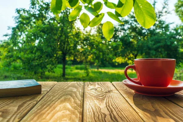 Orange cup of hot coffee and a book on a wooden table in a green garden in the morning. Summer background.