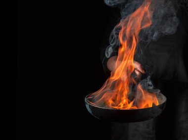 Profesyonel aşçı, siyah arka planda ateş alevi olan tavada yemek pişiriyor. Restoran ve otel servisi konsepti. Ücretsiz reklam alanı