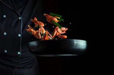 Aşçı ya da şef sebzeli tavada karides pişirir. Deniz ürünleri pişirmek, sağlıklı vejetaryen yiyecekler ve karanlık arka planda yemek. Hareket halindeyken donuyorum. Ücretsiz reklam alanı.