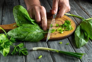 Fresh spinach and green onions are being chopped on a wooden cutting board. The person is preparing for a healthy meal, surrounded by vibrant greens in a cozy kitchen.