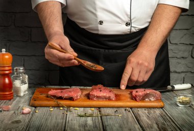 A chef is carefully sprinkling seasoning over three cuts of fresh meat arranged on a wooden cutting board. The setting features a rustic kitchen with stone walls.