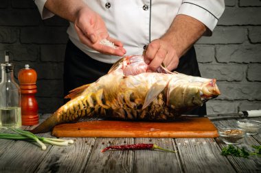 A chef skillfully seasons a freshly caught fish with spices in a rustic kitchen. The wooden table is adorned with ingredients like green onions and chili.