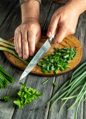 Hands are busy chopping fresh green onions and herbs on a wooden cutting board. The cozy kitchen setting adds warmth to this culinary activity, perfect for meal preparation.