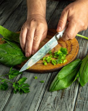 Hands skillfully chop fresh greens and herbs on a wooden cutting board in a warm kitchen. The natural light highlights the vibrant colors of the ingredients.