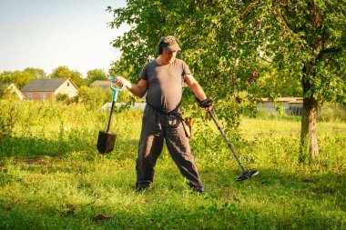A man stands in a vibrant green field, wielding a metal detector in one hand and a small shovel in the other. He searches eagerly, surrounded by trees and houses in late afternoon light.