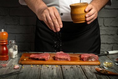 Bir şef, ahşap bir kaptaki tuzu serperek taze eti tatlandırmaya hazırlanır. Köy mutfağında ahşap bir tahta ve çeşitli baharatlar var. Yemek pişirme atmosferini geliştiriyor..