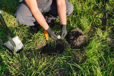 Eldiven takan bir kişi çimenli bir bölgeyi kazarken metal dedektörlü küçük bir nesne tutuyor. Kazmak için aletler güneş ışığı altında yakınlarda yatıyor..