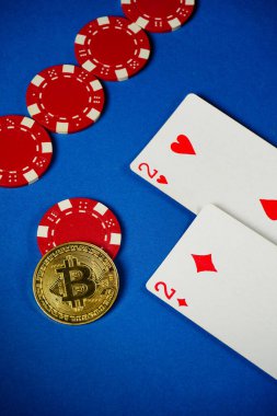 Mavi bir masada, kırmızı poker fişleri iki iskambil kağıdının yanında düzenlenir. Çiplerin arasına bir Bitcoin yerleştirilmiş, geleneksel ve modern oyun karışımı sergileniyor..