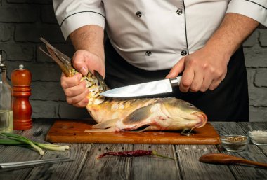 Bir şef, ahşap bir kesme tahtasındaki taze balığı dikkatlice keser. Mutfak ortamında, yakınlardaki malzemelerle bıçak ustalığını gösterir..