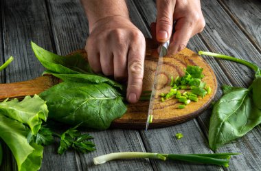 Elleri ustalıkla yeşil soğan ve ıspanak yapraklarını ahşap bir tahtada dilimliyor. Yakınlarda taze bitkiler düzenleniyor, yemek hazırlama sahnesine canlı renkler ekleniyor..