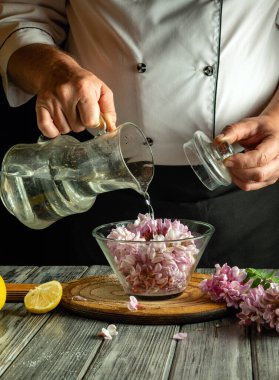 Bir şef dikkatlice bir sürahiden suyu mor çiçeklerle dolu bir kasenin üzerine döker. Taze limon parçaları yakınlarda, kırsal ahşap masa düzenine renk katıyor..
