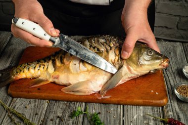 Bir şef, tahta kesme tahtasındaki yeni yakalanan balığı temizlemek için keskin bir bıçak kullanır. Sıcak mutfak atmosferi yakınlardaki çeşitli bitki ve baharatlarla vurgulanır..