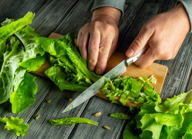 Tahta kesme tahtasının üzerinde taze marul doğrayan biri var. Tahtayı çevreleyen canlı yeşil yapraklar, iyi aydınlatılmış bir mutfakta besleyici bir yemeğin hazırlandığını gösteriyor..