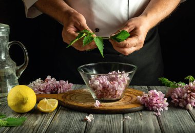 Bir şef cam bir kasede taze otlar ve çiçek yaprakları karışımı dikkatlice hazırlar. Limon dilimleri ve bir sürahi su da kırsal ahşap masanın üzerinde durgun bir pişirme atmosferi yaratıyor..