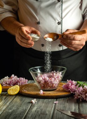 Bir şef ustalıkla narin çiçeklerle dolu bir kaseye şeker döker, tatlı bir tatlı hazırlar. Taze limon dilimleri ve çiçekler ahşap masayı süslüyor, sahneyi güzelleştiriyor..